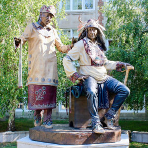 Indigenous Land (1) A photo of a statue featuring two indigenous persons in traditional clothing and attire—a male chief is seated and a clan mother stands. The sculpture is on the campus of Concordia University, Montreal.